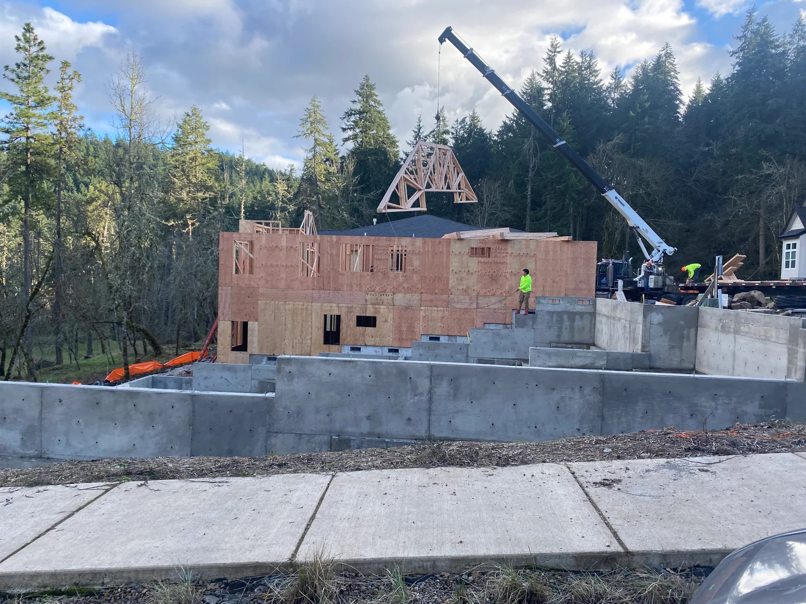 Wood-framed house structure with concrete foundation, roof trusses, and construction crane on wooded site