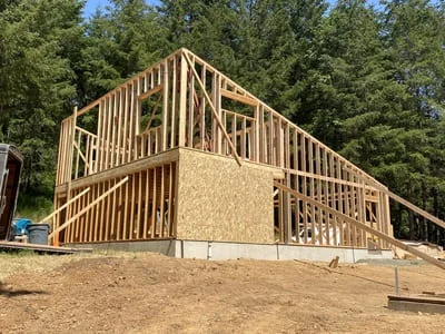 Two-story home framing with lumber studs, roof trusses, and OSB sheathing on bare ground